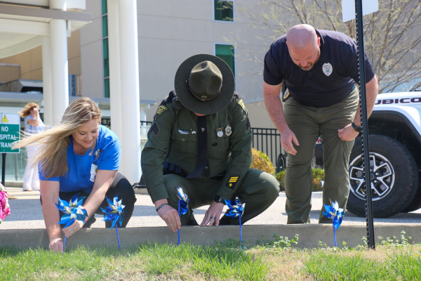Three people, a woman in a blue T-shirt, a state trooper in his army green uniform and a police officer in black place blue and silver pinwheels in the ground.