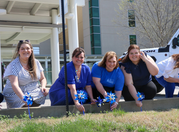 Four white women kneel in front of grass with blue and silver pinwheels in their hands. 
